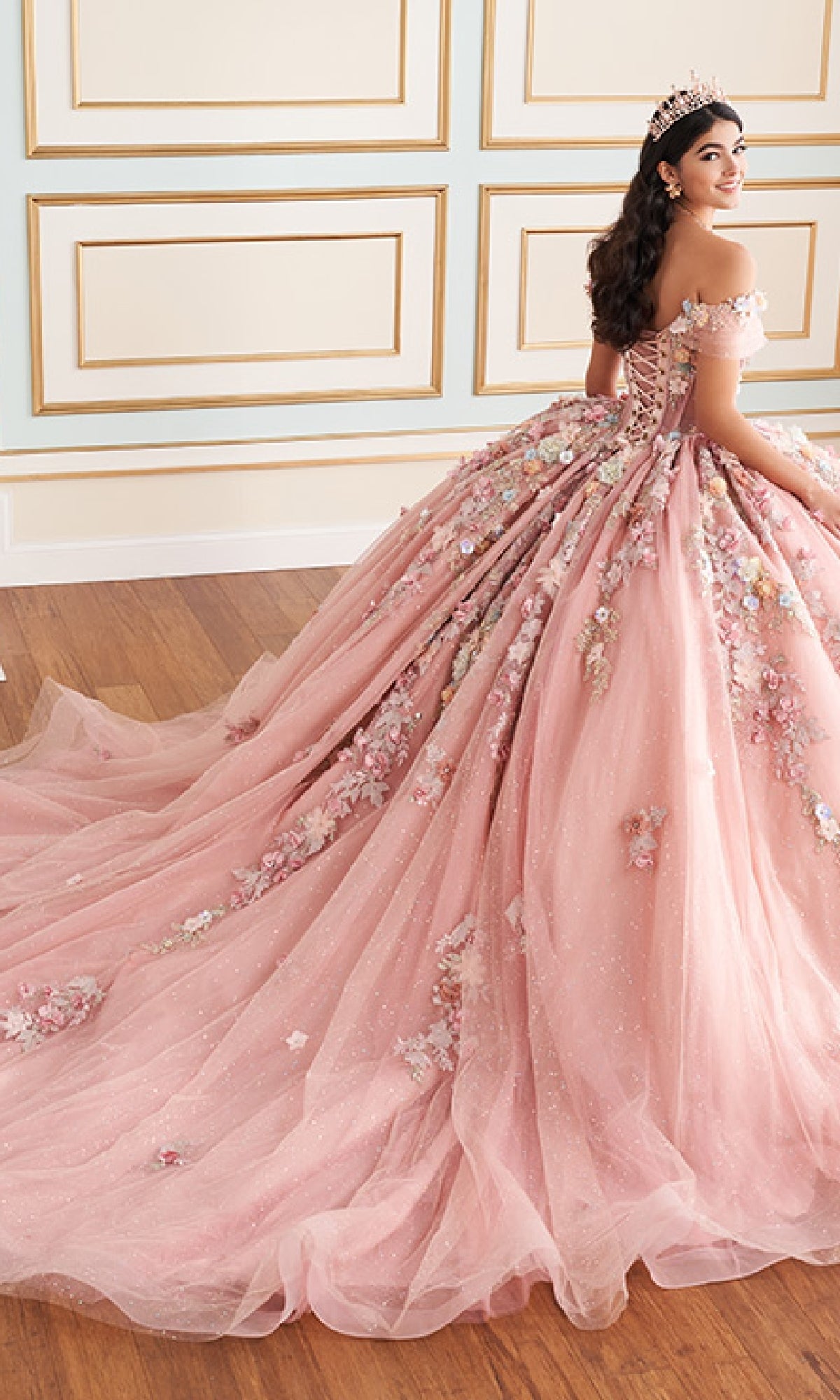 A young woman in the Quinceanera Dress PR30175 By Princesa, featuring off-shoulder 3D pink florals and delicate beading, wears a tiara as she sits in an elegant room with paneled walls and wooden floors.